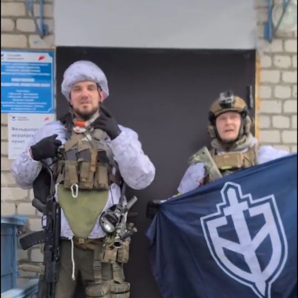 Ein etwas verpixelt Screenshot zeigt Denis Nikitin in Kampfmontur. Nikitin gestikuliert in Richtung Kamera. Neben ihm hält ein Mann - ebenfalls in Militärkleidung - ein blaues Banner mit dem Logo des Russischen Freiwilligenkorps hoch.