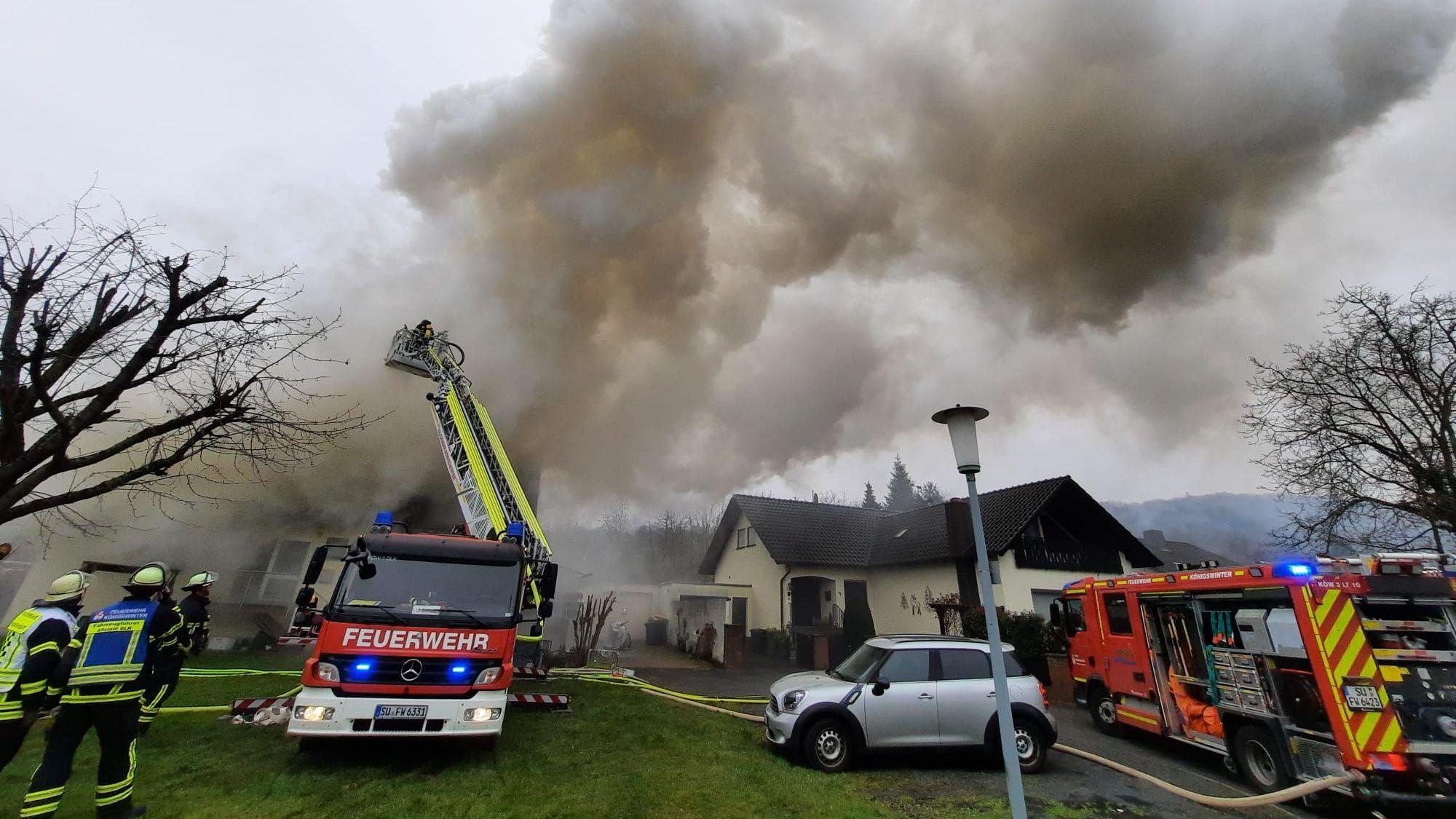 Über einem Haus steigt eine dichte, graue Rauchwolke auf. Von einer Drehleiter aus löschen Feuerwehrleute den brennenden Dachstuhl..