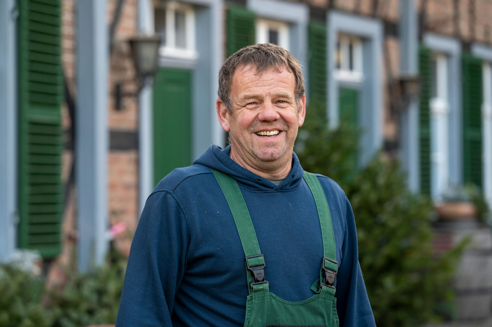 Landwirt Hans-Gerd Eyberg trägt einen blauen Hoodie und eine grüne Latzhose und lacht in die Kamera.