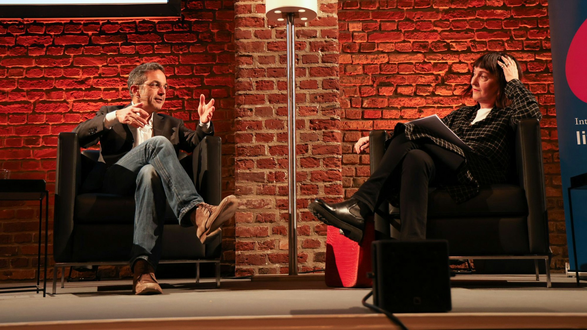 Navid Kermani und Sonia Mickich sitzen auf der Bühne in den Balloni Hallen in Köln-Ehrenfeld. Sie schauen einander an, sind in einem Gespräch. Kermani gestikuliert, Mickich hat die Hand am Kopf und hört ihm zu.