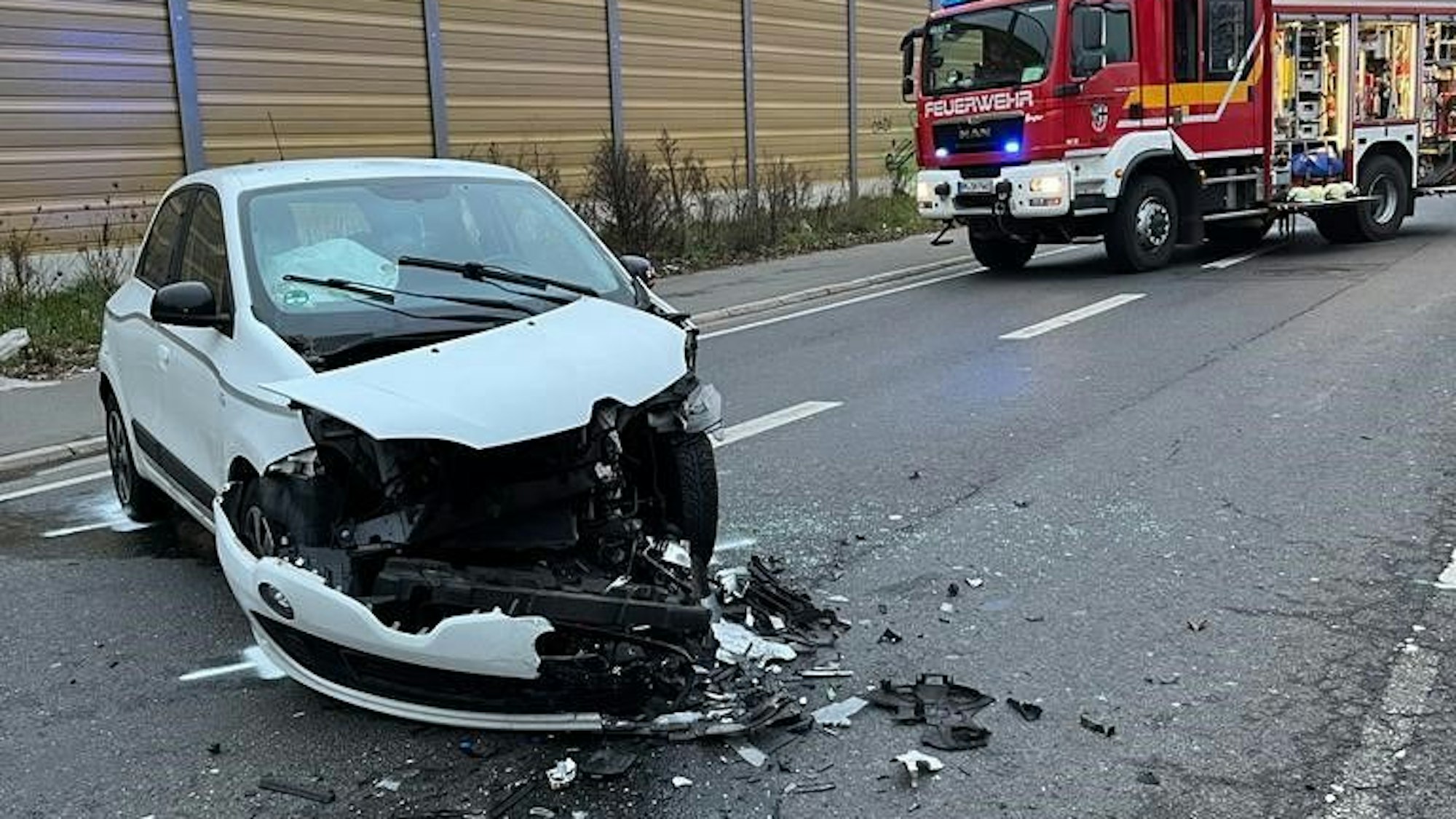 Ein Auto mit kaputter Front steht auf einer Straße. Im Hintergrund steht ein Einsatzfahrzeug der Feuerwehr.