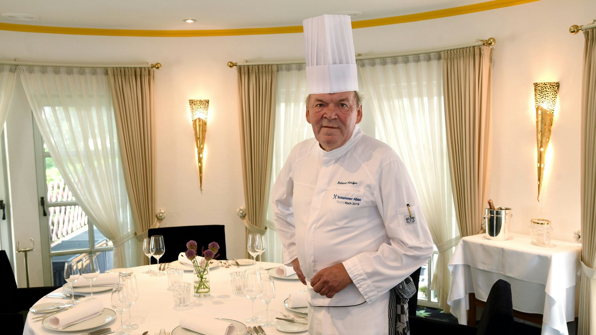 Maitre Erhard Schäfer trägt eine Kochmütze und steht an einem festlich gedeckten Tisch.