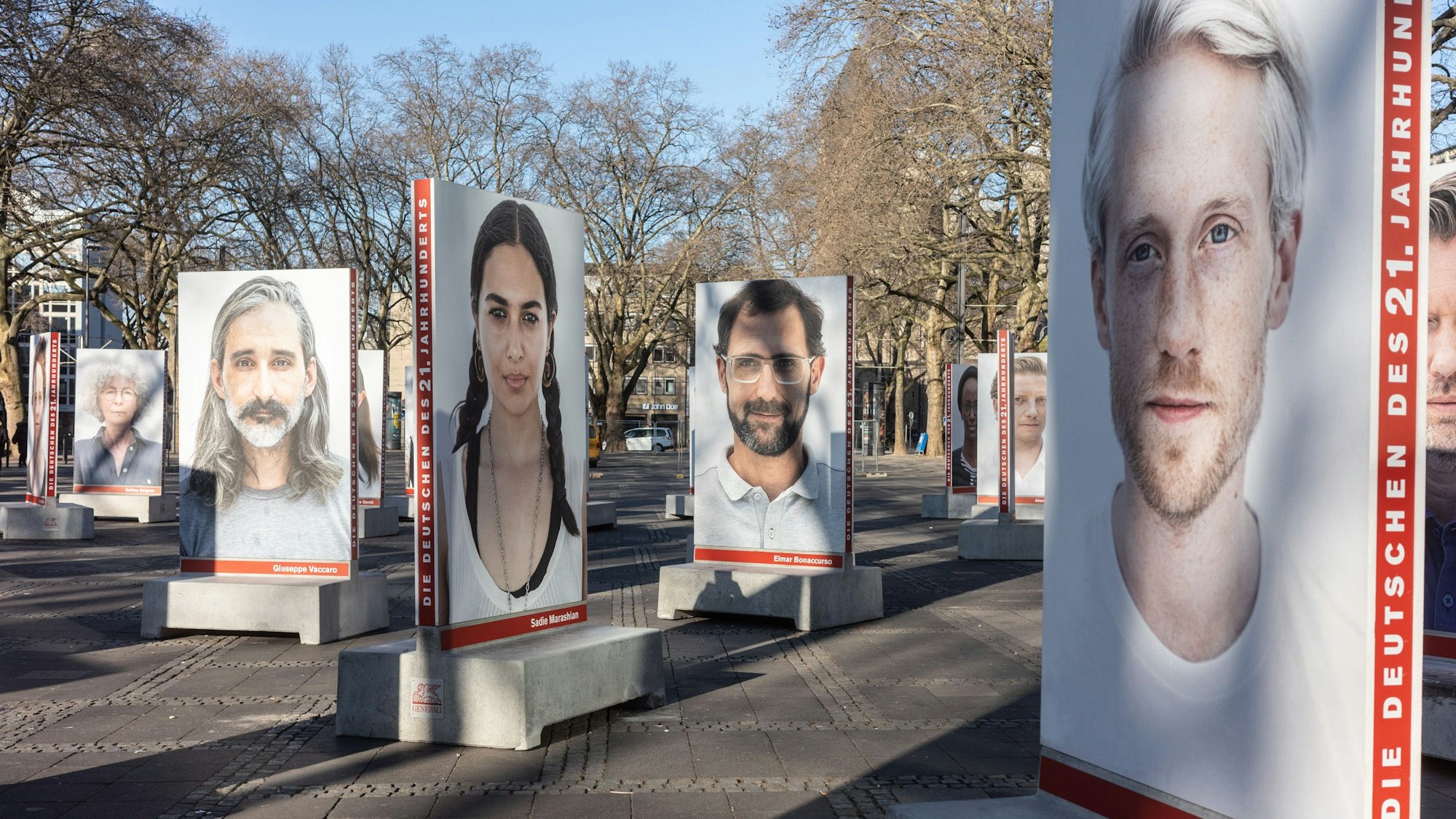 Auf dem Kölner Neumarkt stehen große Betonsockel, auf denen Portraits von diversen Menschen sind, die in die Kamera schauen. Die Sockel dienen unten auch als Bänke.