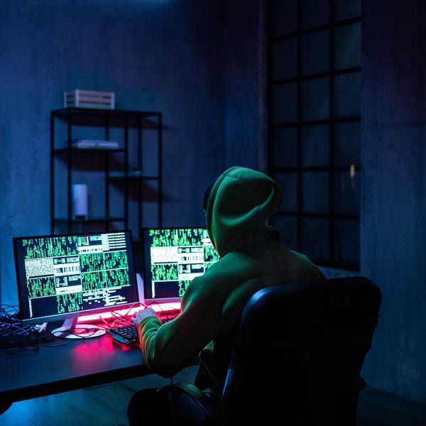 Ein Hacker mit Kapuze sitzt mit dem Rücken zur Kamera an einem Tisch. Das Licht ist ausgeschaltet, nur auf dem Bildschirm leuchtet es geheimnisvoll.