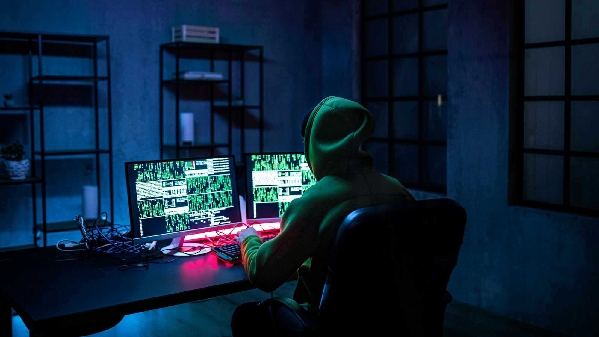 Ein Hacker mit Kapuze sitzt mit dem Rücken zur Kamera an einem Tisch. Das Licht ist ausgeschaltet, nur auf dem Bildschirm leuchtet es geheimnisvoll.