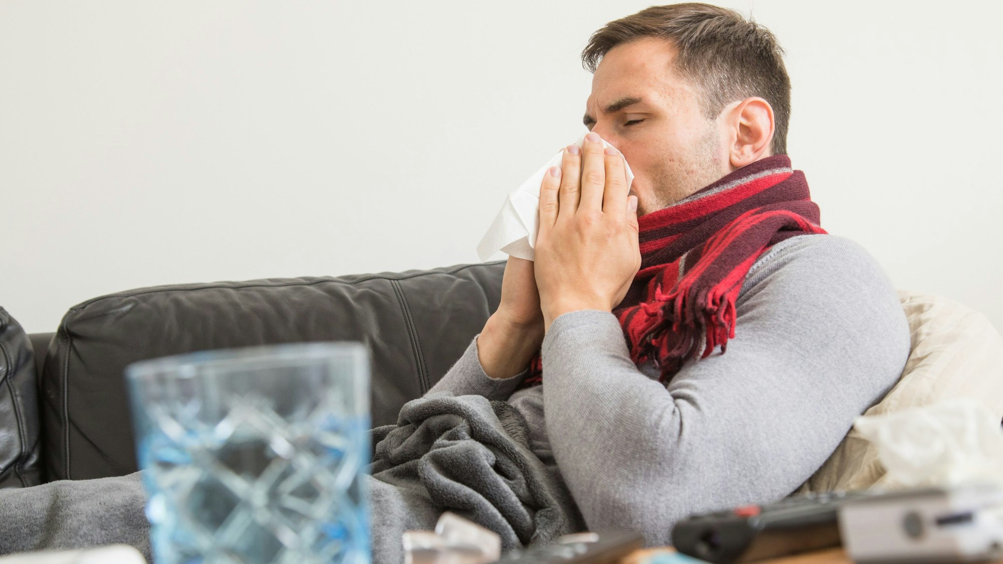 Ein Mann sitzt erkältet auf dem Sofa und putzt sich die Nase.