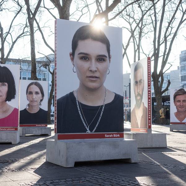 Auf dem Kölner Neumarkt stehen große Betonsockel, auf denen Porträts von diversen Menschen sind, die in die Kamera schauen. Die Sockel dienen unten auch als Bänke. Hinter einem Porträt strahlt die Sonne.