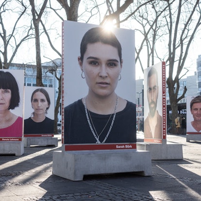 Auf dem Kölner Neumarkt stehen große Betonsockel, auf denen Porträts von diversen Menschen sind, die in die Kamera schauen. Die Sockel dienen unten auch als Bänke. Hinter einem Porträt strahlt die Sonne.
