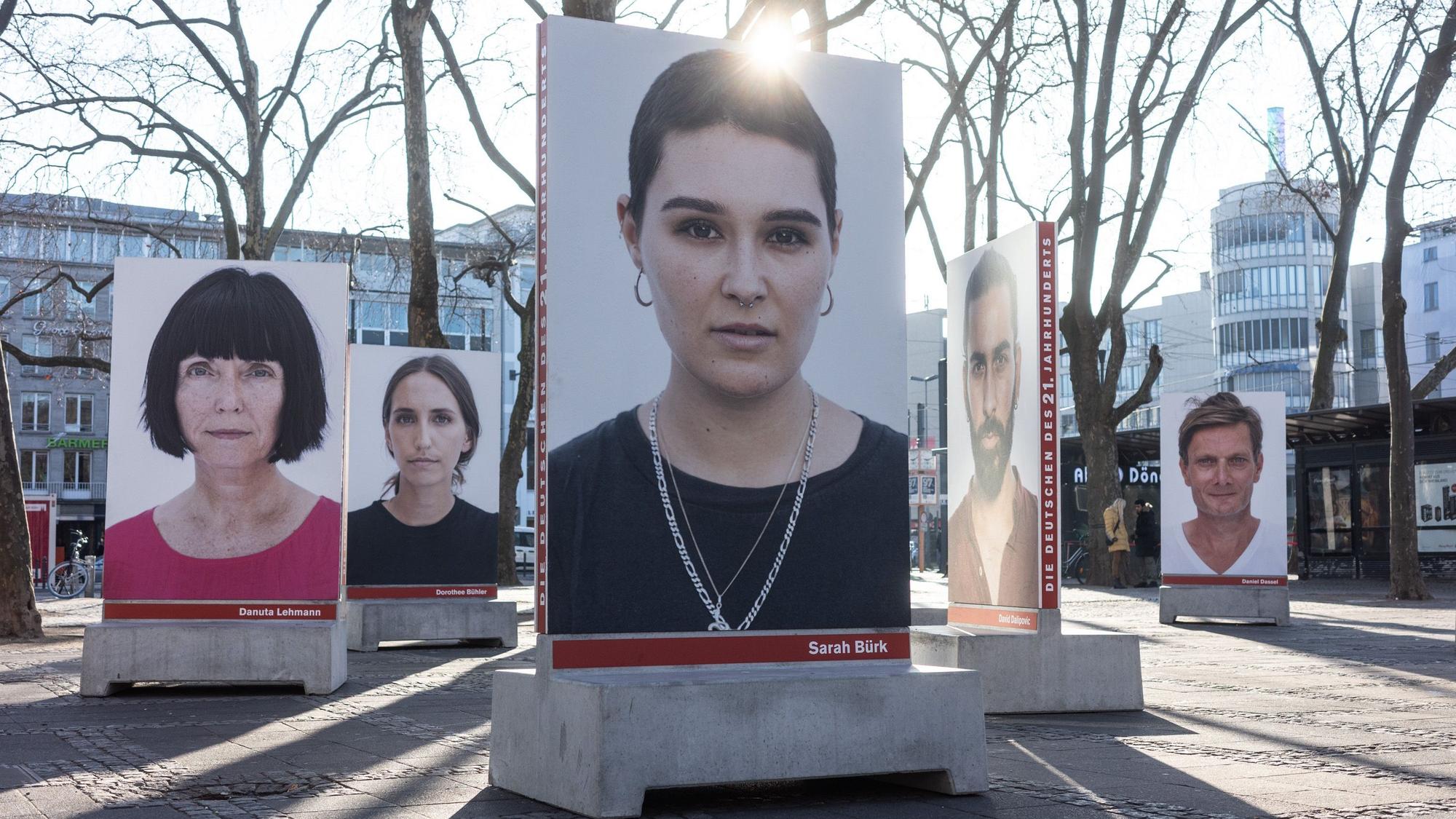 Auf dem Kölner Neumarkt stehen große Betonsockel, auf denen Porträts von diversen Menschen sind, die in die Kamera schauen. Die Sockel dienen unten auch als Bänke. Hinter einem Porträt strahlt die Sonne.