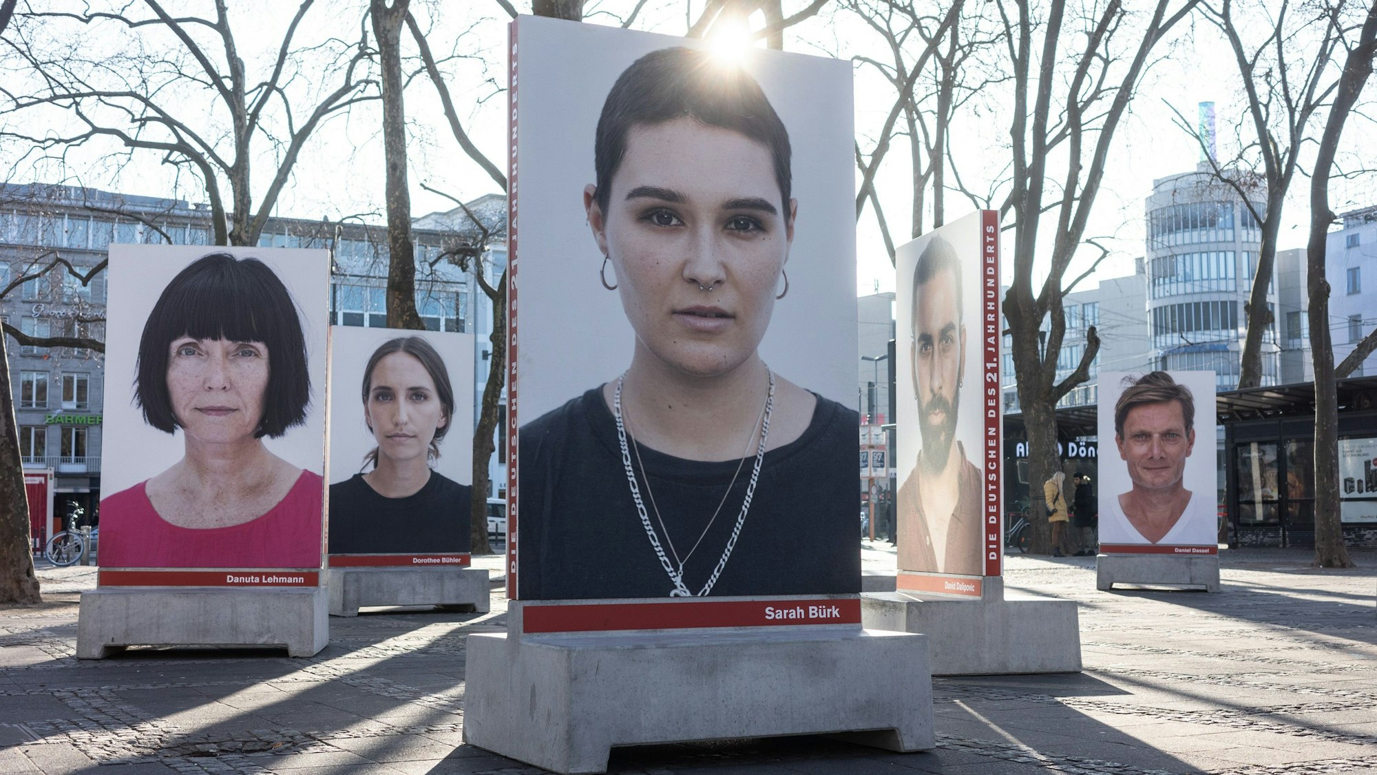 Auf dem Kölner Neumarkt stehen große Betonsockel, auf denen Porträts von diversen Menschen sind, die in die Kamera schauen. Die Sockel dienen unten auch als Bänke. Hinter einem Porträt strahlt die Sonne.