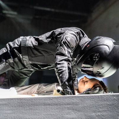 Der Schauspieler Justus Maier beugt sich im schwarzen Motrorrad-Anzug mit Helm über eine Statistin. Szene aus Robert Borgmanns „Meta-Sleep“, eine musiktheatrale Installation nach Motiven von »Leonce und Lena« am Schauspiel Köln.