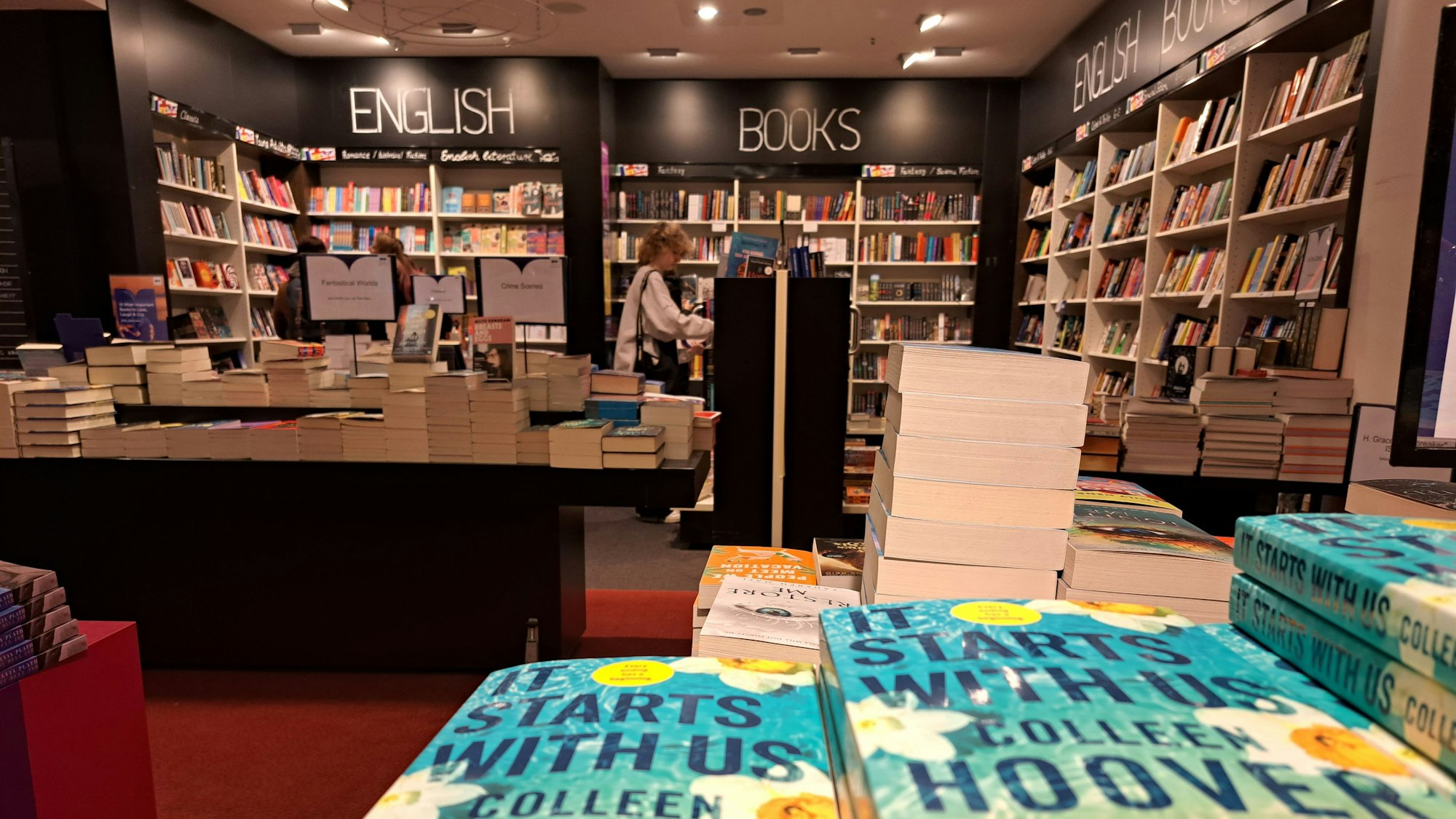 Blick auf die Abteilung für englische Bücher in der Mayersche-Buchhandlung am Neumarkt in der Kölner Innenstadt. Im Vordergrund liegen einige Ausgaben von Colleen Hoovers Taschenbuch „It starts with us“. Hinten sind vier große Wandregale mit der Überschrift "English Books". Davor ist ein weiteres Regal sowie mehrere Thementische, auf denen sich die Bücher stapeln.