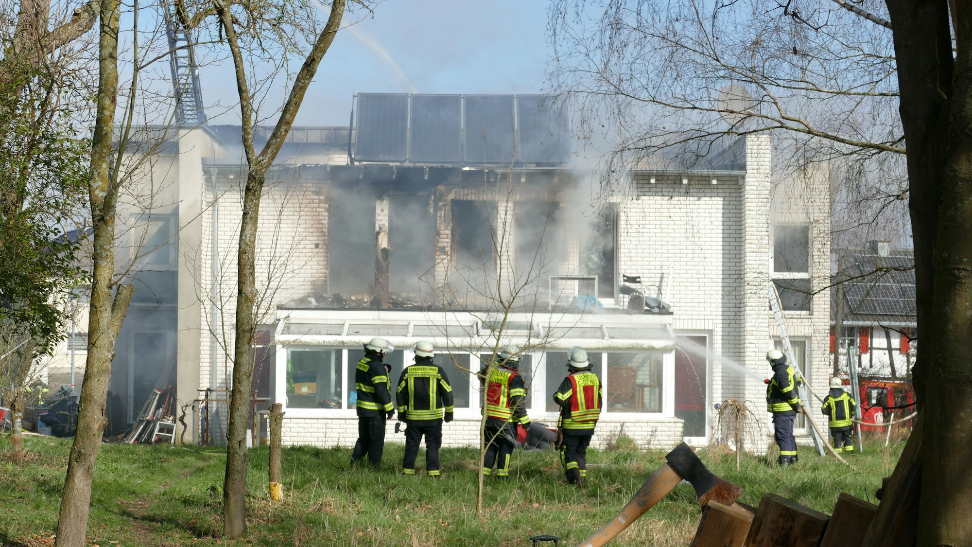 Feuerwehrleute löschen ein Feuer in einem weißen Haus. Sie stehen in einem Garten.