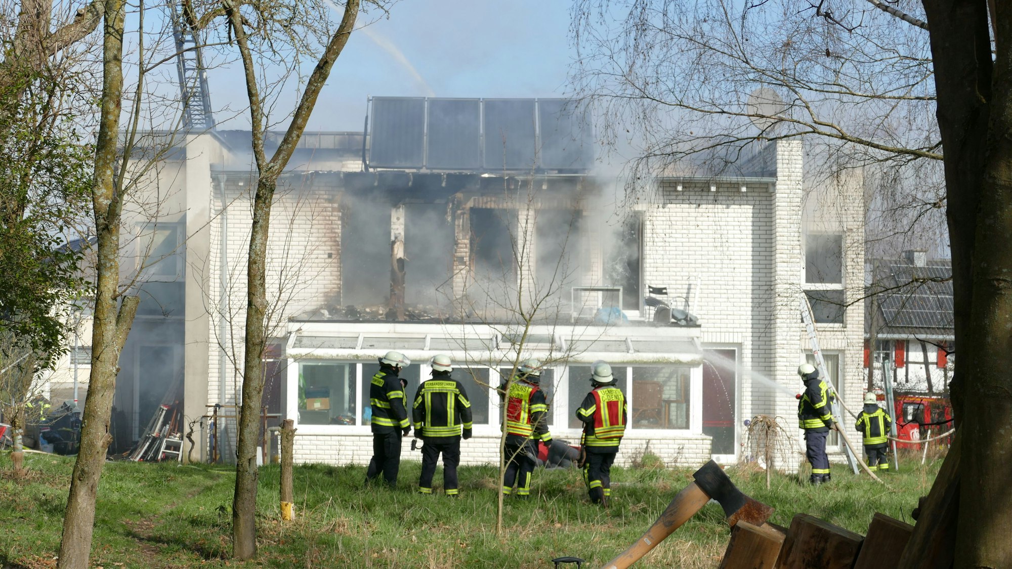 Feuerwehrleute löschen ein Feuer in einem weißen Haus. Sie stehen in einem Garten.
