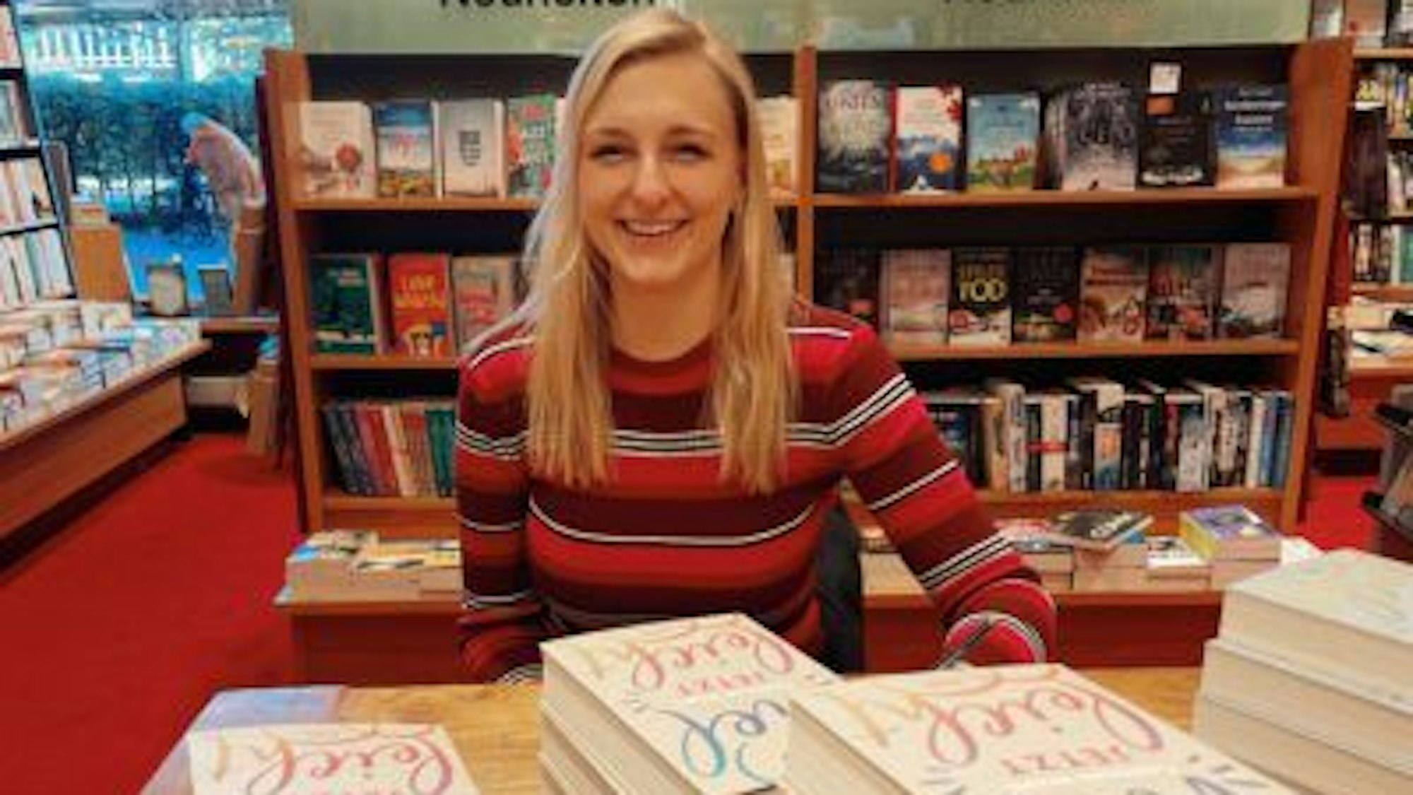 Die Autorin Carolin Wahl sitzt in einer Buchhandlung und lächelt in die Kamera. Vor ihr liegen Ausgaben ihres Buchs „Vielleicht jetzt“.