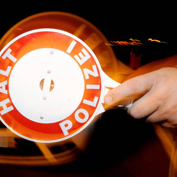 Ein Polizist hält nachts ein Fahrzeug an. Zu sehen ist im Vordergrund eine Polizeikelle mit der Aufschrift „Halt, Polizei“.