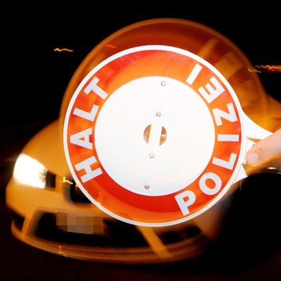 Ein Polizist hält nachts ein Fahrzeug an. Zu sehen ist im Vordergrund eine Polizeikelle mit der Aufschrift „Halt, Polizei“.
