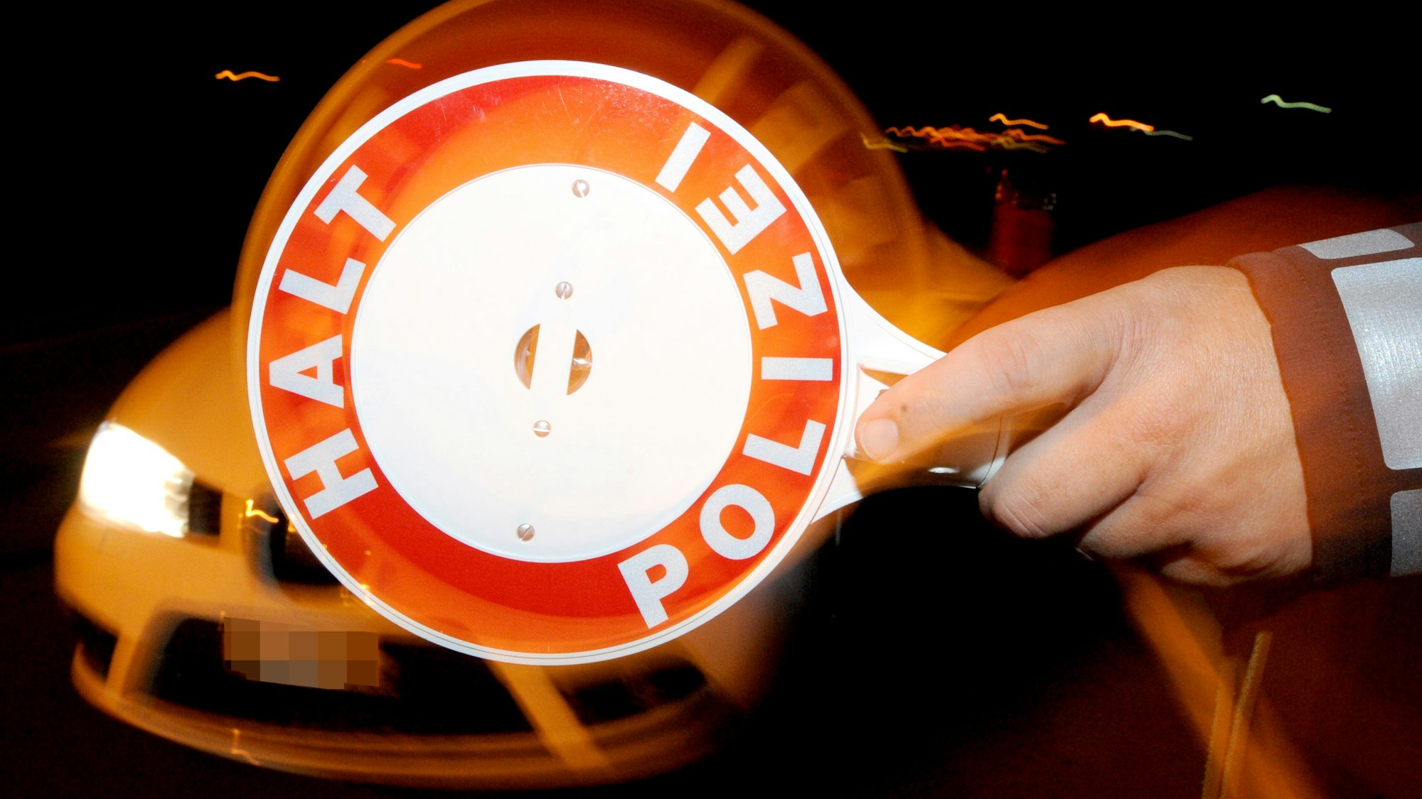 Ein Polizist hält nachts ein Fahrzeug an. Zu sehen ist im Vordergrund eine Polizeikelle mit der Aufschrift „Halt, Polizei“.