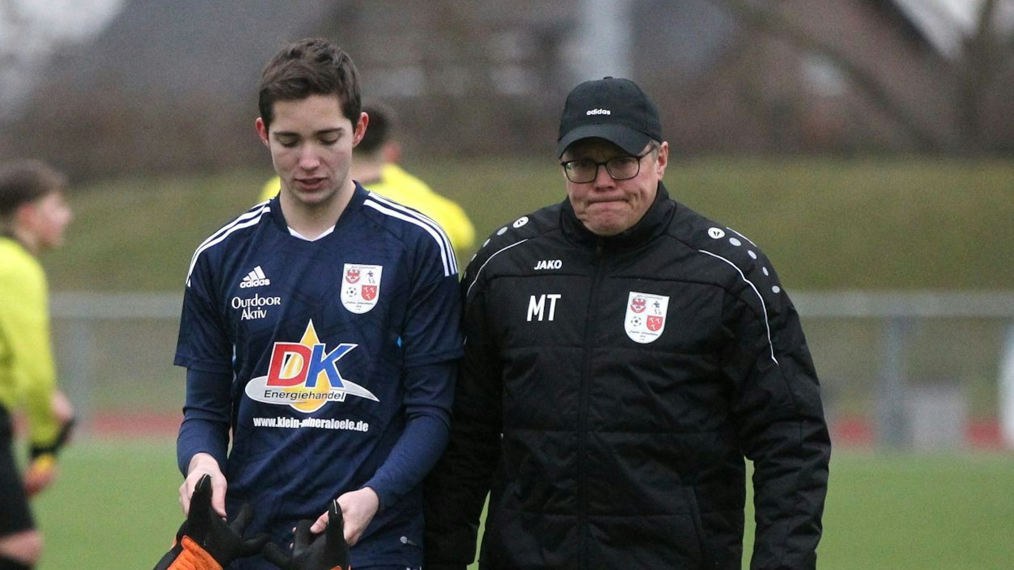 Ein Fußballtrainer schaut missmutig in die Kamera. Neben ihm geht ein Spieler.
