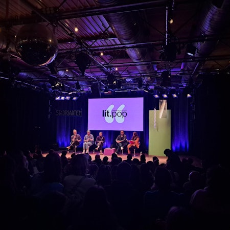 Valentin Moritz (links), Philipp Winkler, Joris Bas Backer, Dinçer Güçyeter und Mithu Sanyal bei der lit.pop im Kölner Stadtgarten am 4. März. Sie sitzen in der Reihe auf der Bühne. Hinter ihnen ist das lilane Logo der lit.pop groß auf einem Bildschirm. Vor ihnen sitzt das Publikum.