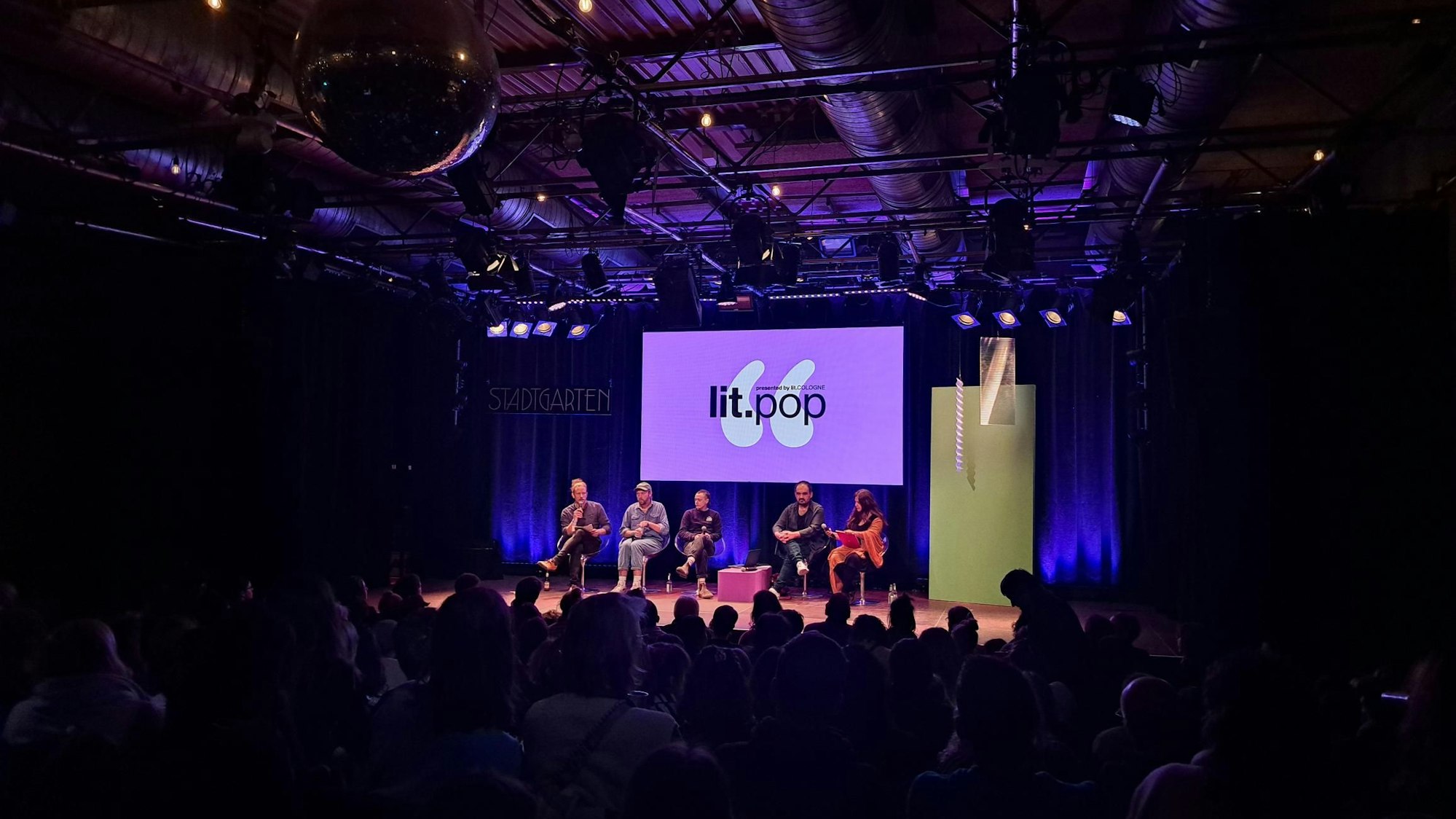 Valentin Moritz (links), Philipp Winkler, Joris Bas Backer, Dinçer Güçyeter und Mithu Sanyal bei der lit.pop im Kölner Stadtgarten am 4. März. Sie sitzen in der Reihe auf der Bühne. Hinter ihnen ist das lilane Logo der lit.pop groß auf einem Bildschirm. Vor ihnen sitzt das Publikum.