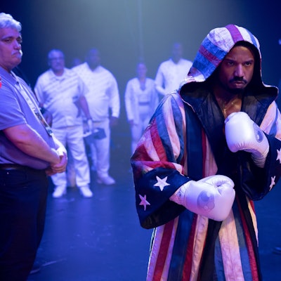 Ein Mann mit Boxhandschuhen und Mantel geht an vielen Menschen vorbei.
