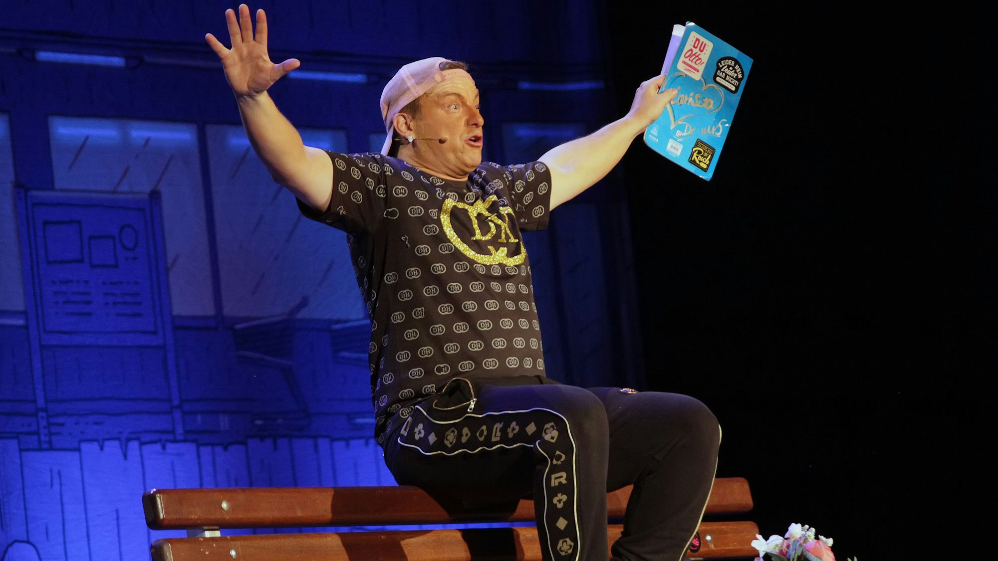 Der Comedian sitzt auf der Bühne auf einer Bank und hebt die Hände nach oben.