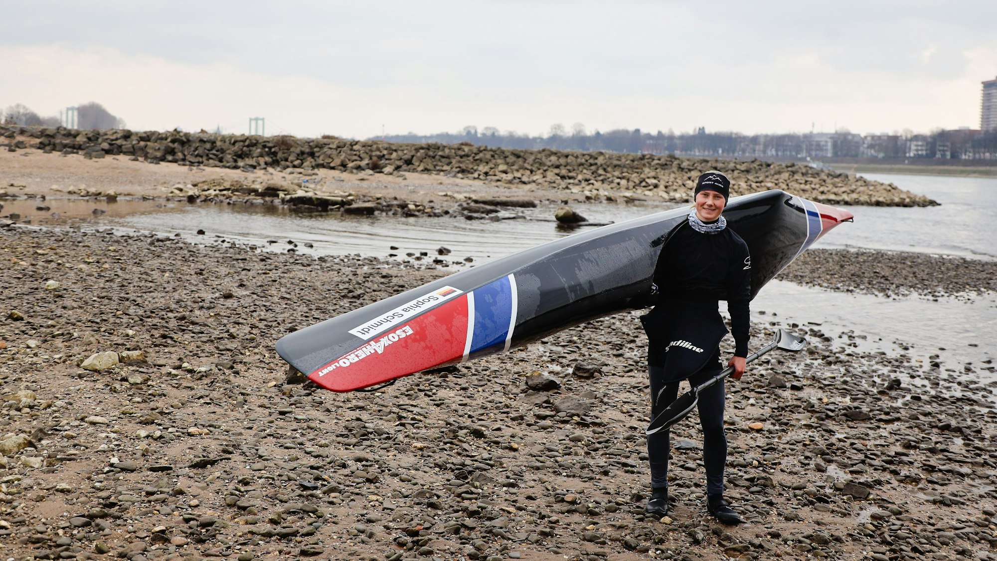 Wildwasser-Sportlerin Sophia Schmidt steht mit ihrem Boot auf der Schulter am Rheinufer in Poll. Der Fluss führt wenig Wasser, man sieht Sandbänke und Steinbuhnen.