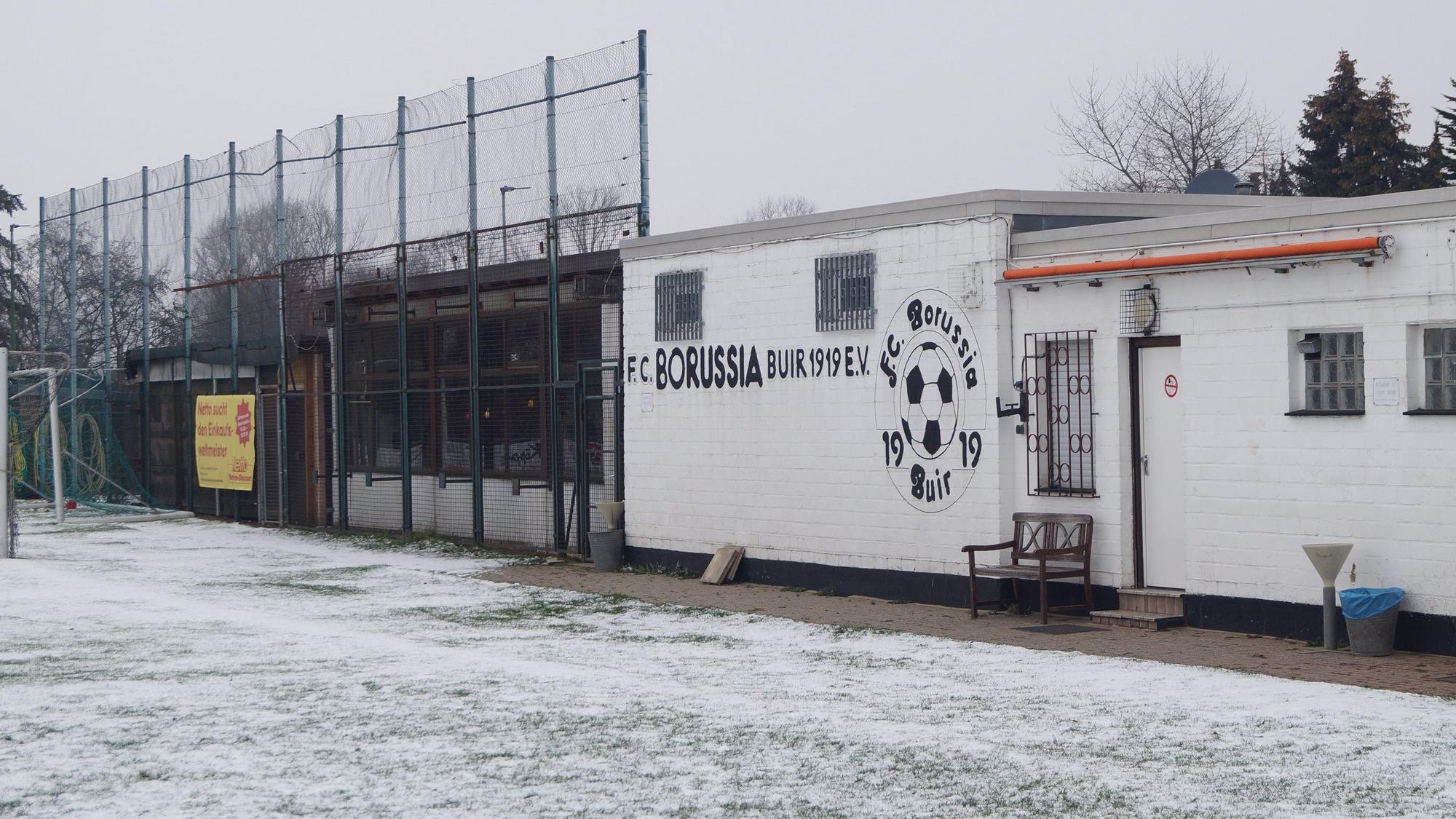Der Rasen ist mit Schnee bedeckt, dahinter steht ein einstöckiges Vereinsheim mit der Aufschrift „FC Borussia Buir“.