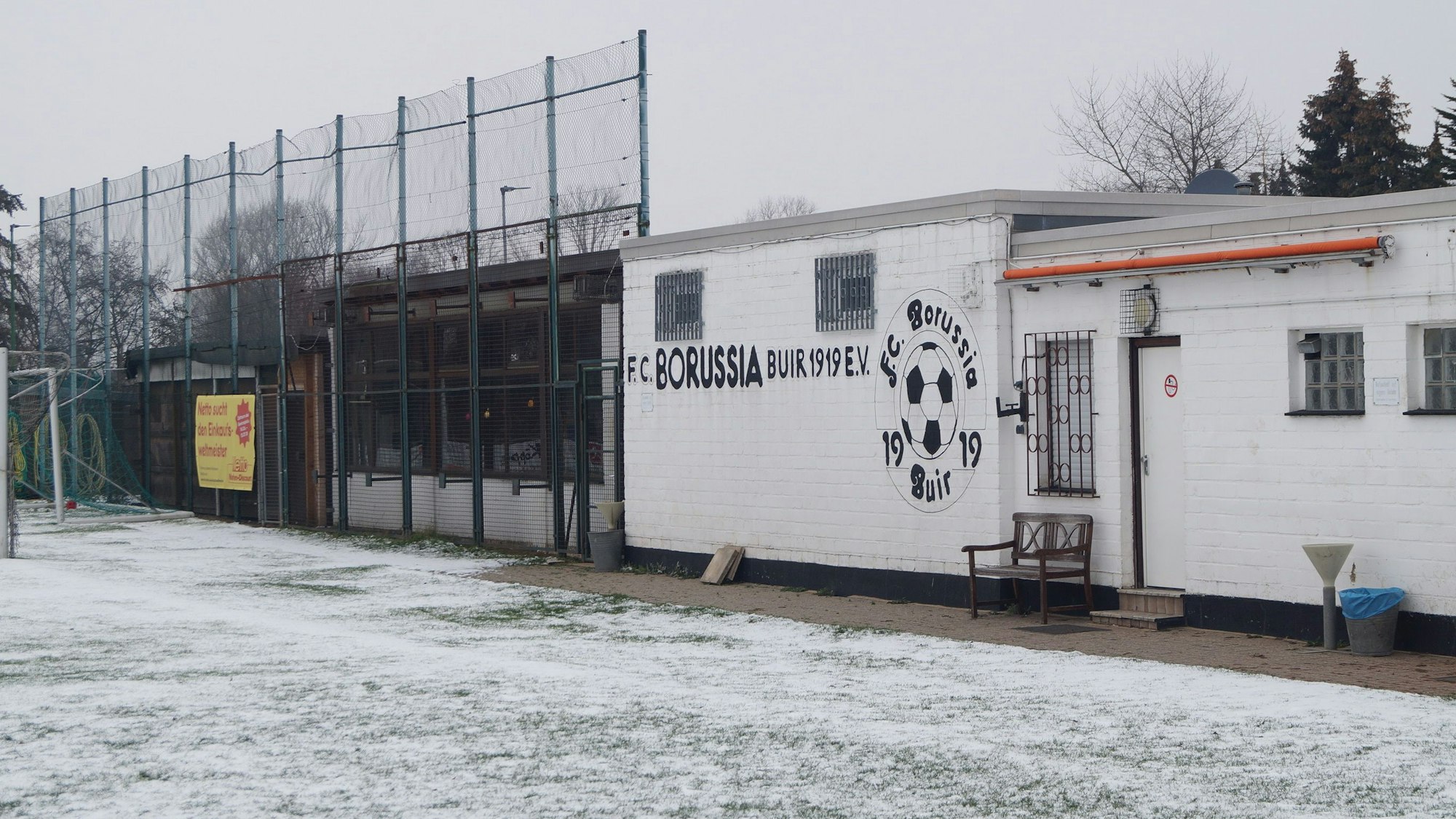 Der Rasen ist mit Schnee bedeckt, dahinter steht ein einstöckiges Vereinsheim mit der Aufschrift „FC Borussia Buir“.