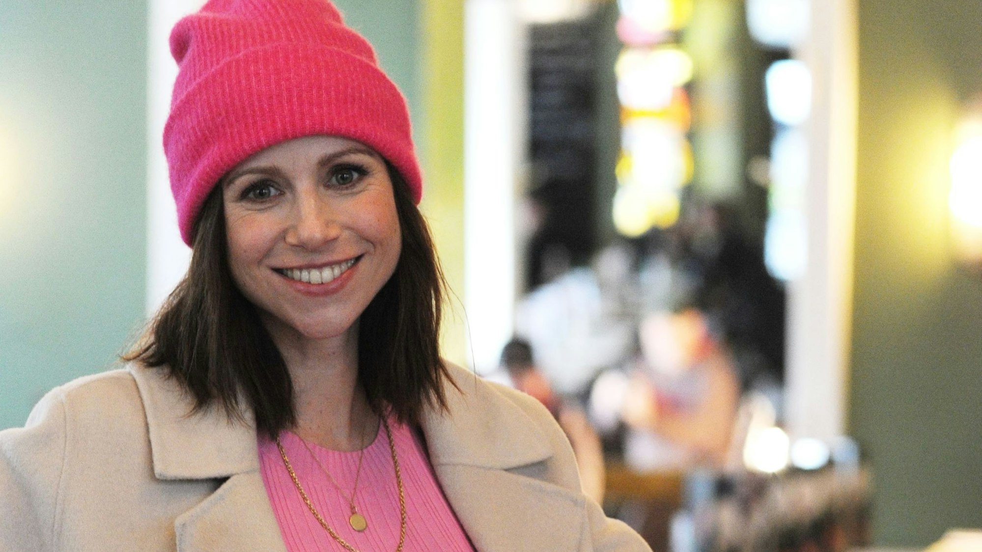 Die Schauspielerin Katrin Heß sitzt am Tisch und trinkt Kaffee, sie trägt eine pinke Mütze und ein pinkes Kleid.