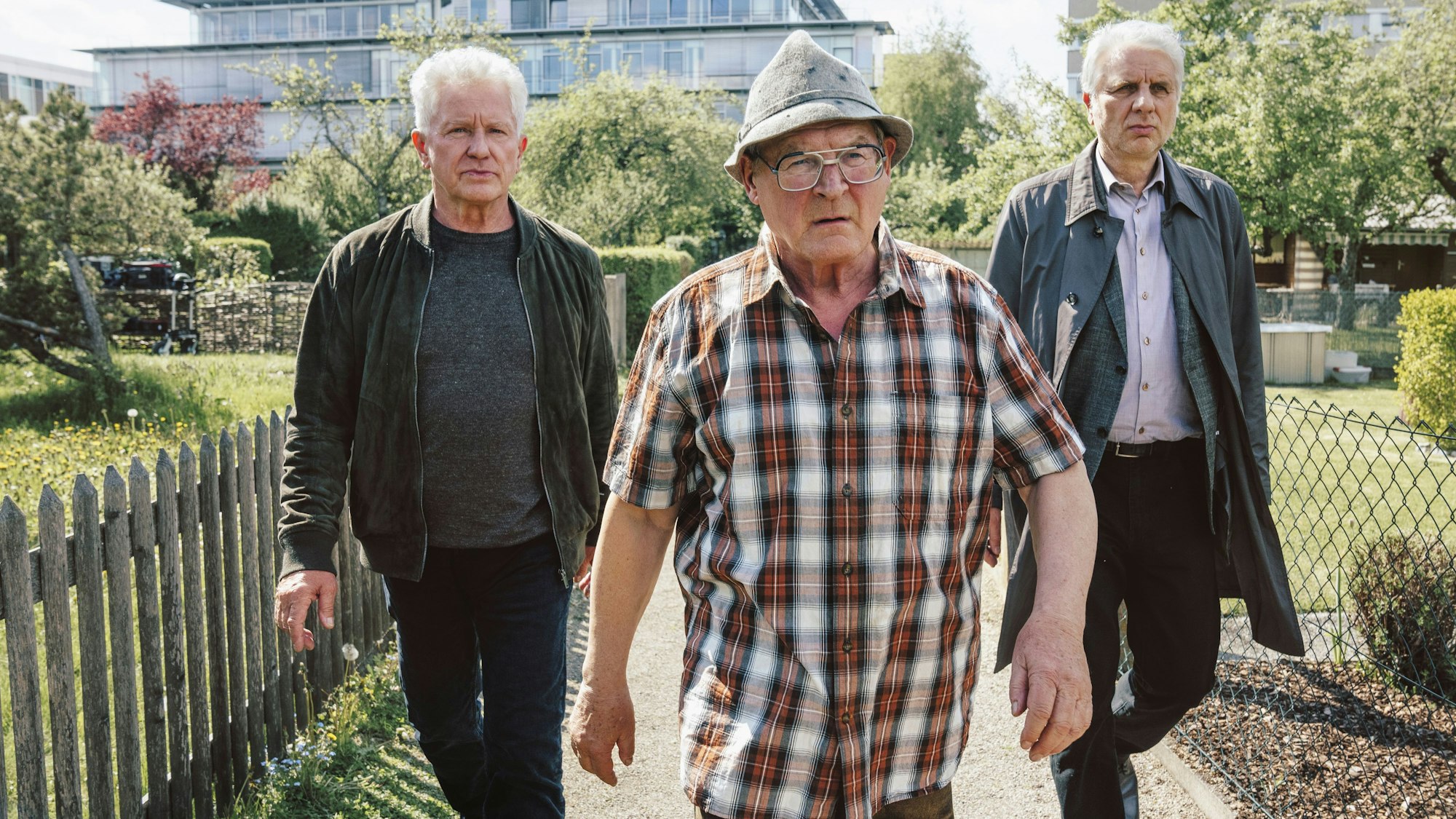 Ivo Batic (Miroslav Nemec, links) und Franz Leitmayr (Udo Wachtveitl, rechts) gehen mit dem Verdächtigen Hackl (Burghart Klaußner) durch eine Kleingartensiedlung. Batic und Leitmeyr schauen ernst, fast genervt. Hackl sieht wütend aus und geht vor ihnen.