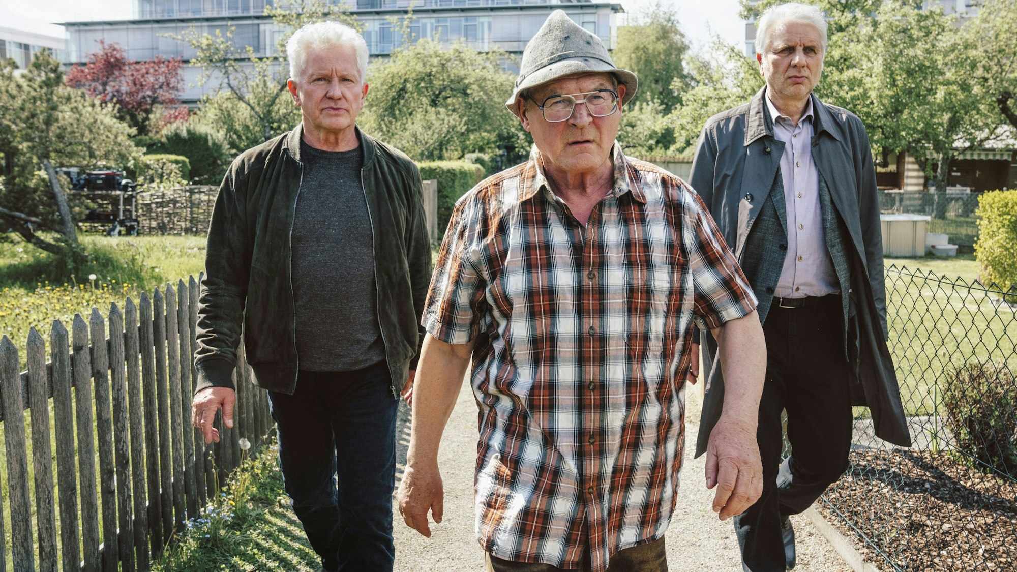 Ivo Batic (Miroslav Nemec, links) und Franz Leitmayr (Udo Wachtveitl, rechts) gehen mit dem Verdächtigen Hackl (Burghart Klaußner) durch eine Kleingartensiedlung. Batic und Leitmeyr schauen ernst, fast genervt. Hackl sieht wütend aus und geht vor ihnen.