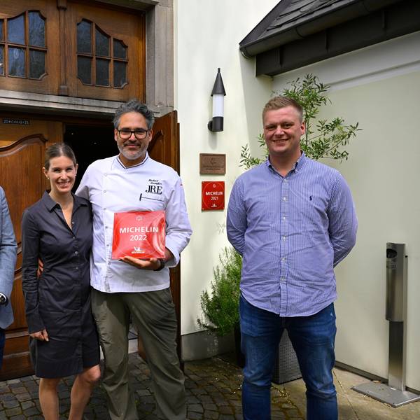 Vier Personen stehen vor einem Gebäude mit einer großen Holztüre. Ein Mann hält eine rote Plakette mit der Michelin-Auszeichnung in der Hand.