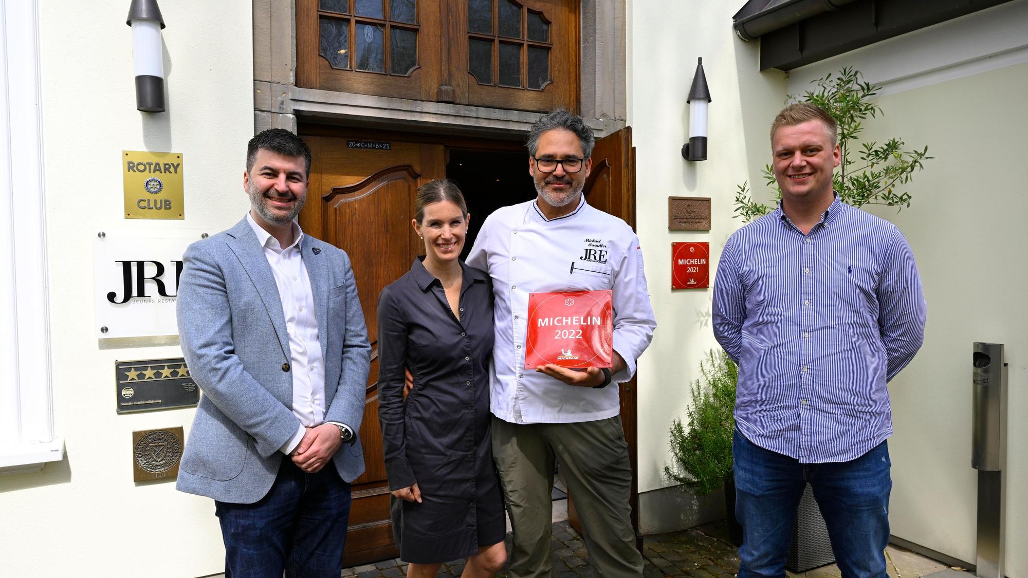 Vier Personen stehen vor einem Gebäude mit einer großen Holztüre. Ein Mann hält eine rote Plakette mit der Michelin-Auszeichnung in der Hand.