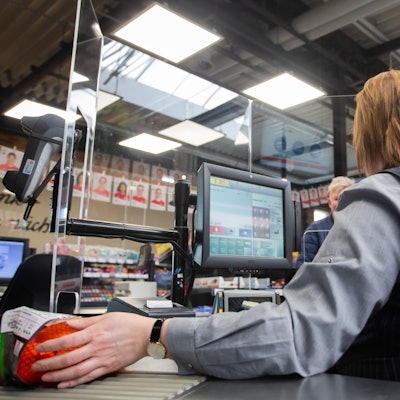 An einer Kasse in einem Supermarkt wird die Kassiererin mit einer Plexiglasscheibe, einem sogenannten "Spuckschutz", geschützt-