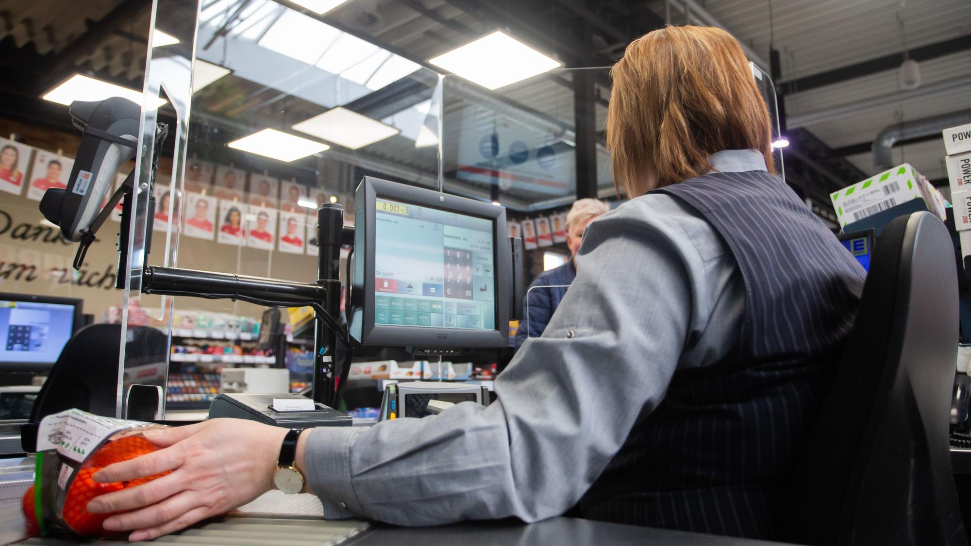An einer Kasse in einem Supermarkt wird die Kassiererin mit einer Plexiglasscheibe, einem sogenannten "Spuckschutz", geschützt-