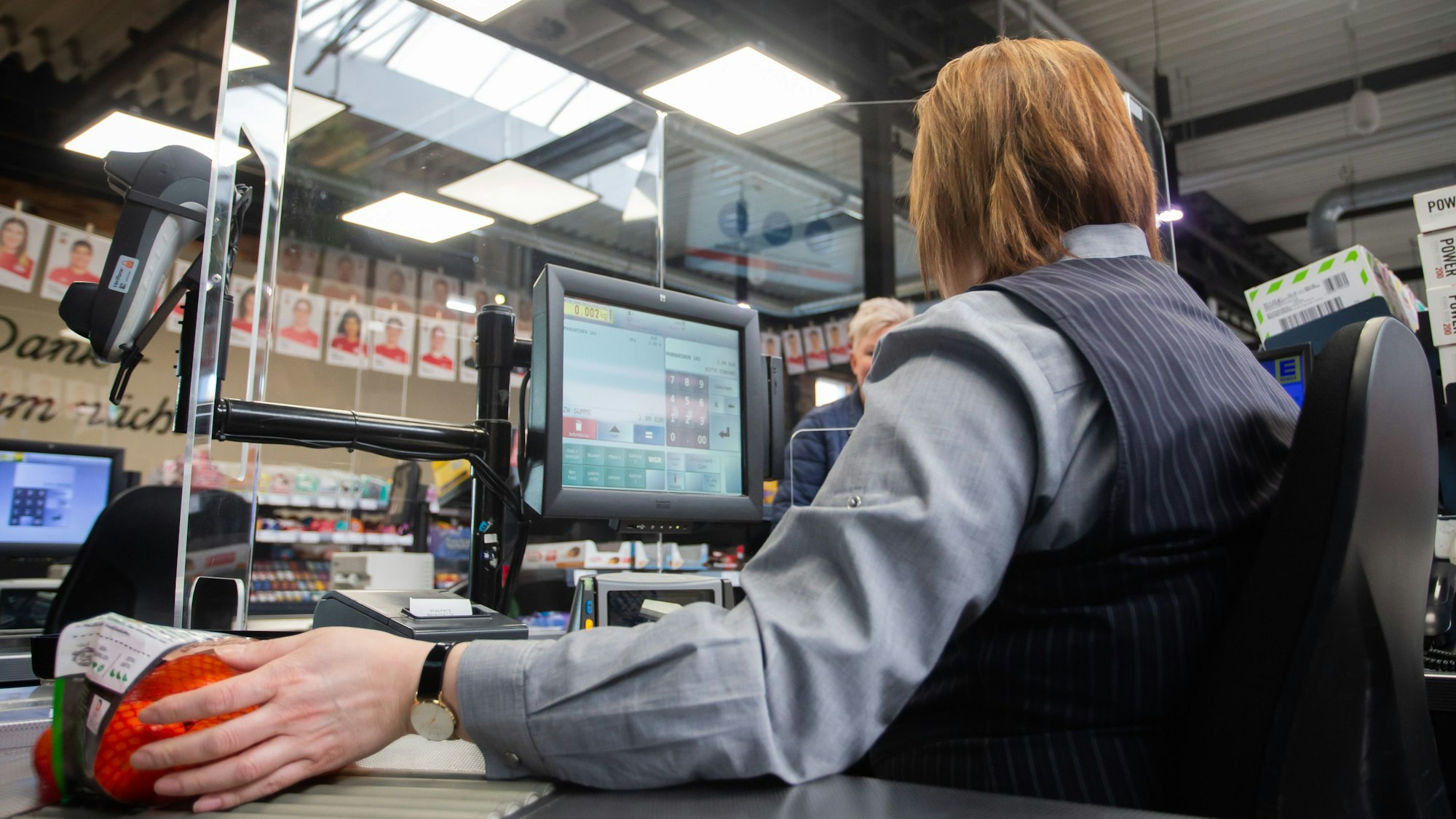 An einer Kasse in einem Supermarkt wird die Kassiererin mit einer Plexiglasscheibe, einem sogenannten "Spuckschutz", geschützt-