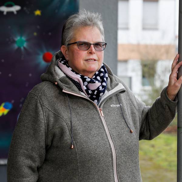 Katharina H. ist im Porträt zu sehen. Sie steht vor einer mit Weltraummotiven bemalten Wand und stützt eine Hand an auf einen Pfosten. Sie trägt ein gepunktetes Halstuch und eine getönte Brille. Ihr Haar ist grau und kurz geschnitten.