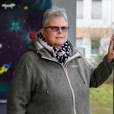 Katharina H. ist im Porträt zu sehen. Sie steht vor einer mit Weltraummotiven bemalten Wand und stützt eine Hand an auf einen Pfosten. Sie trägt ein gepunktetes Halstuch und eine getönte Brille. Ihr Haar ist grau und kurz geschnitten.