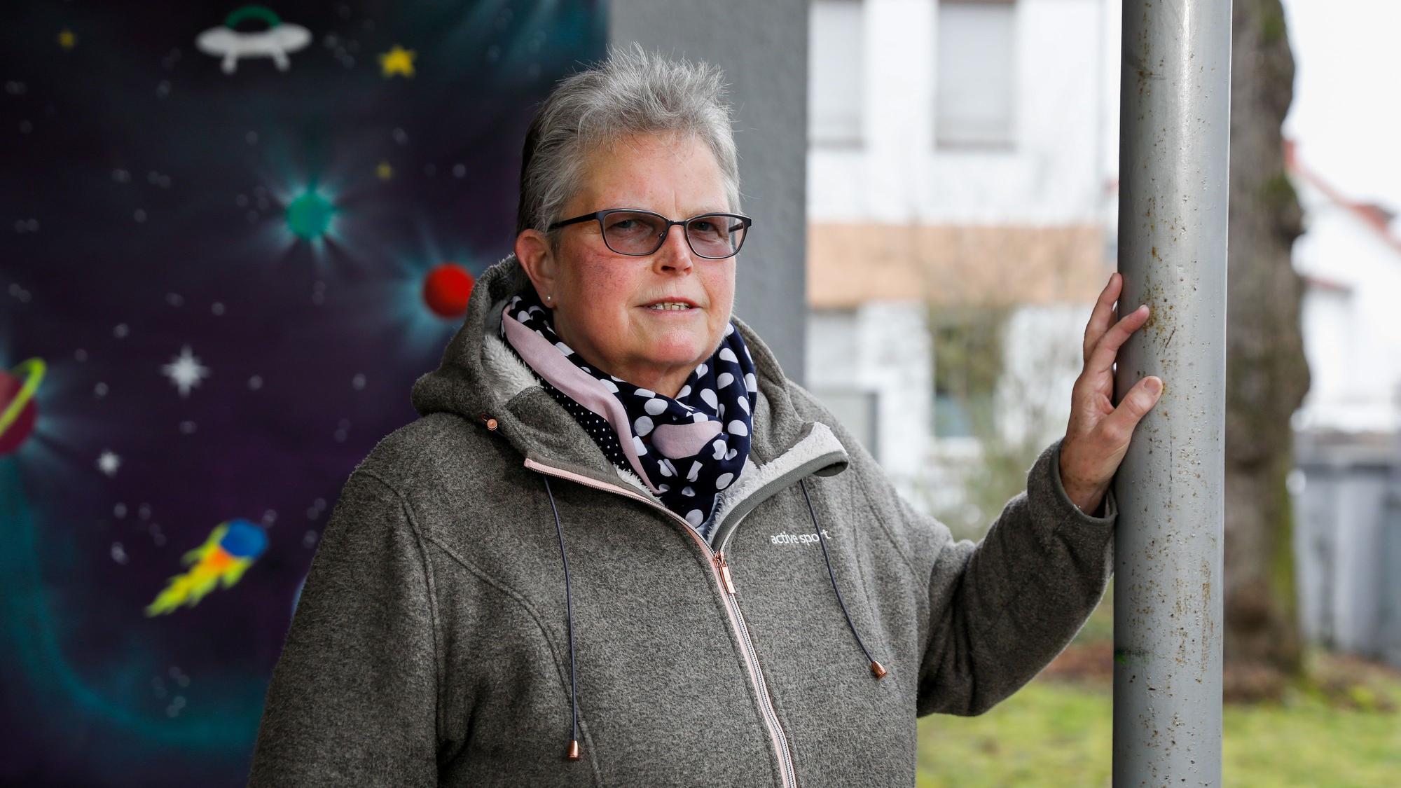 Katharina H. ist im Porträt zu sehen. Sie steht vor einer mit Weltraummotiven bemalten Wand und stützt eine Hand an auf einen Pfosten. Sie trägt ein gepunktetes Halstuch und eine getönte Brille. Ihr Haar ist grau und kurz geschnitten.