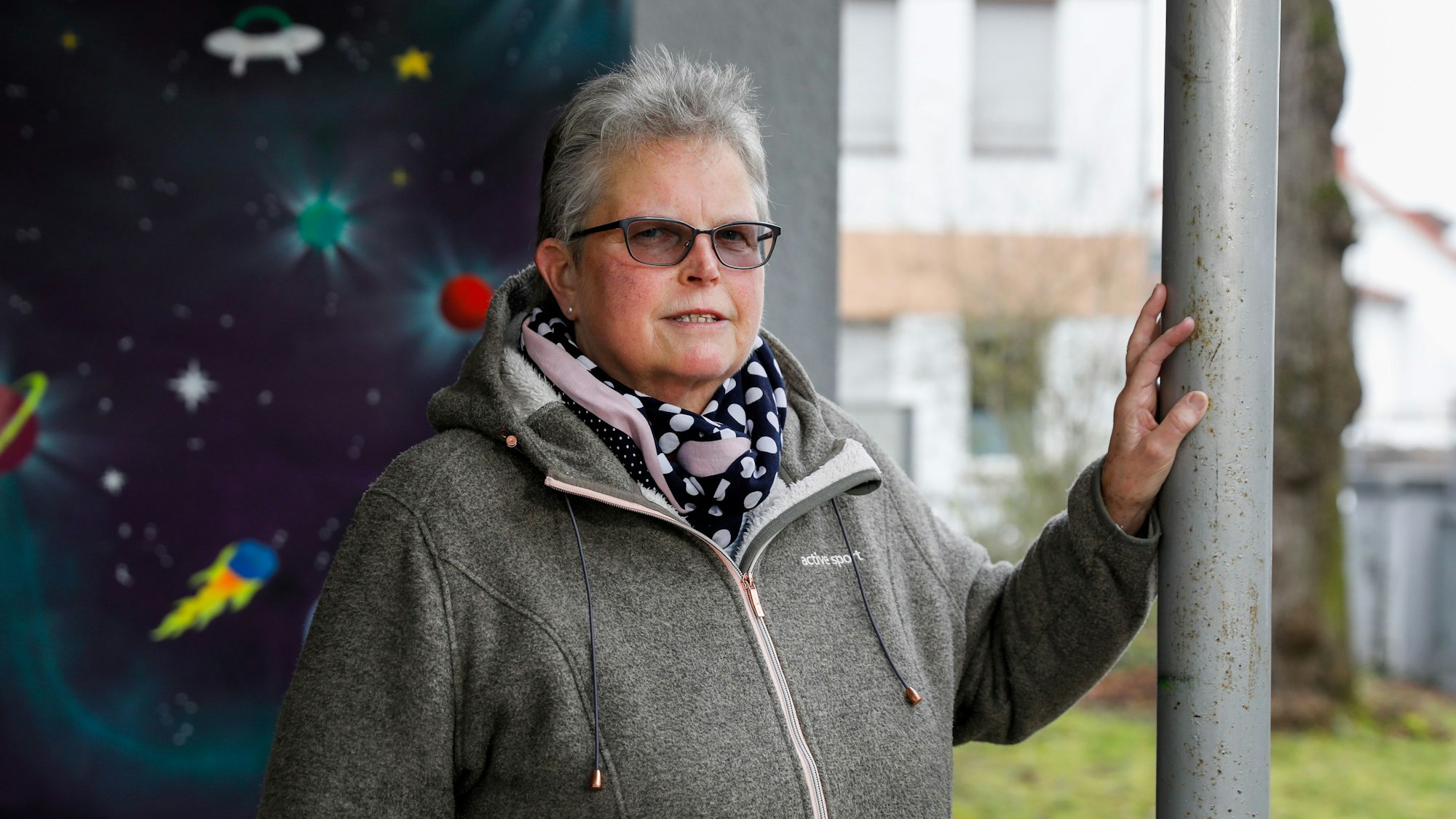 Katharina H. ist im Porträt zu sehen. Sie steht vor einer mit Weltraummotiven bemalten Wand und stützt eine Hand an auf einen Pfosten. Sie trägt ein gepunktetes Halstuch und eine getönte Brille. Ihr Haar ist grau und kurz geschnitten.