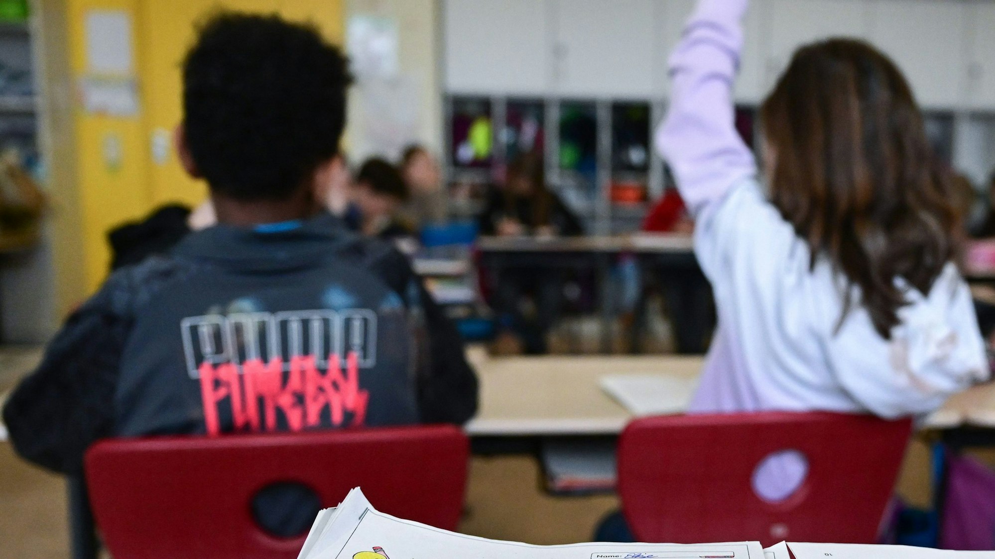 Zwei Grundschüler sitzen in einer Grundschulklasse. Eine Schülerin hebt den Finger.