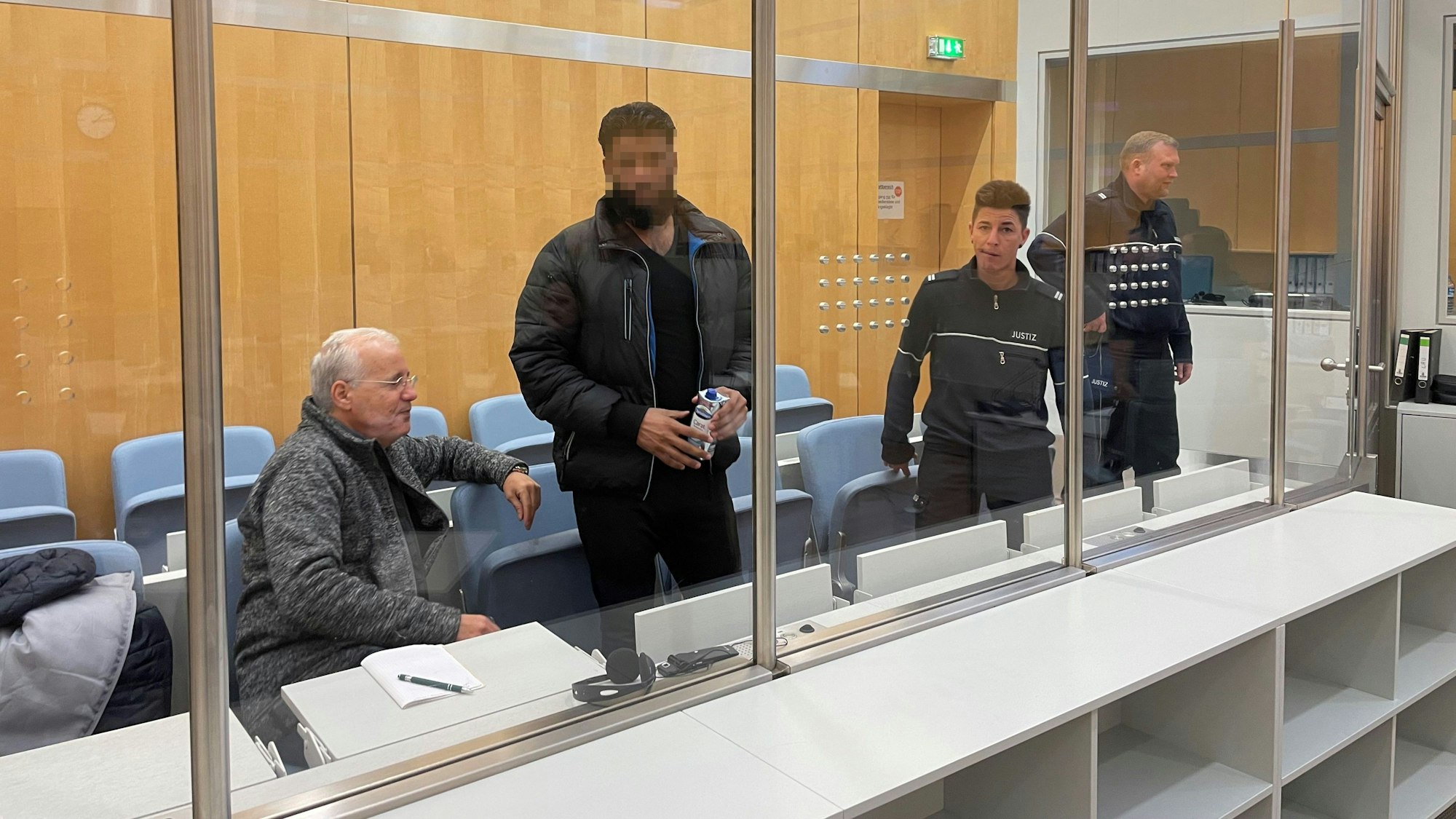 Khaled A. steht im Gericht vor der Anklagebank. Er hat einen Tetrapack Wasser in der Hand, sein Gesicht ist gepixelt.
