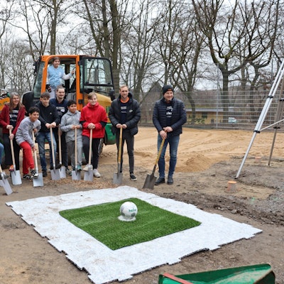 Marcel Risse mit Kindern und Schaufel in der Hand vor dem Kunstrasen, dahinter steht ein Bagger