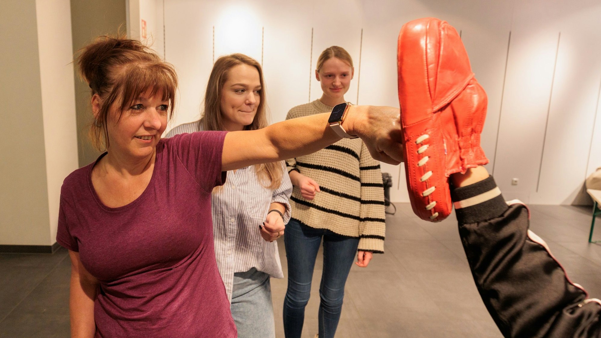 Eine Frau schlägt auf einen Boxhandschuh. Im Hintergrund stehen zwei weitere junge Frauen.
