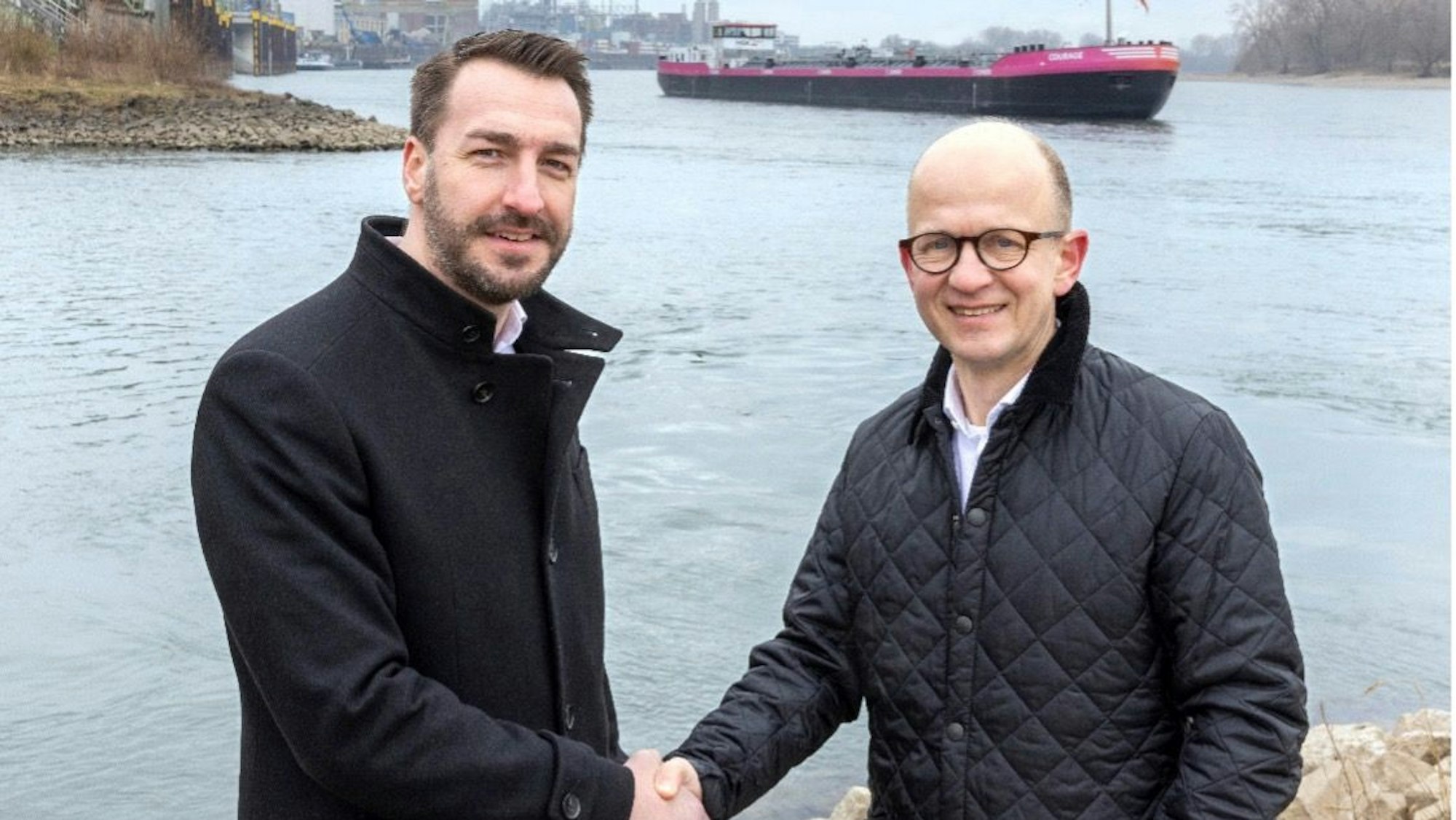 Steffen Bauer (links), Chef von HGK Shipping, steht mit Hanno Brümmer, dem Leiter von Covestros Liefergeschäft in Europa, am Rhein. Im Hintergrund das Schiff „Courage“