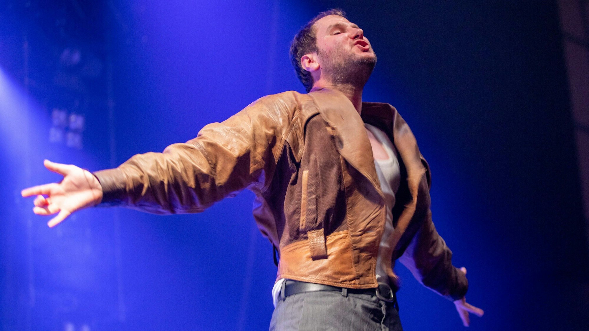 Sänger Marco Michael Wanda von der Band Wanda im Konzert in der Innsbrucker Olympiahalle im Juni 2022. Er hat die Arme aus- und die Brust vorgestreckt und trägt seine abgewetzte braune Lederjacke.