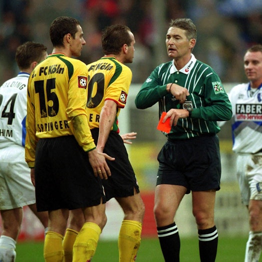 Zwei Spieler des 1. FC Köln in gelben Trikots diskutieren mit dem Schiedsrichter, der eine rote Karte in der Hand hält.