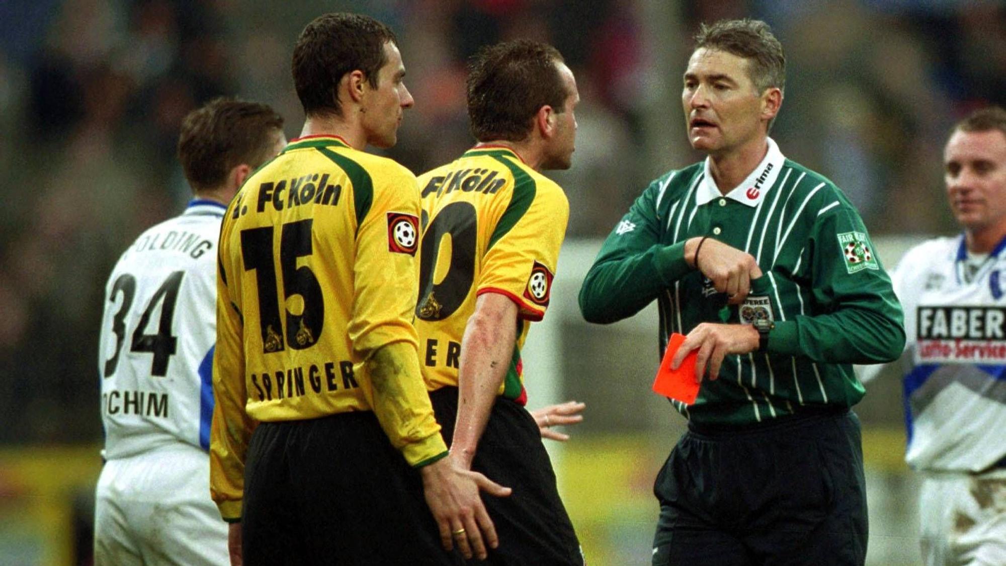 Zwei Spieler des 1. FC Köln in gelben Trikots diskutieren mit dem Schiedsrichter, der eine rote Karte in der Hand hält.